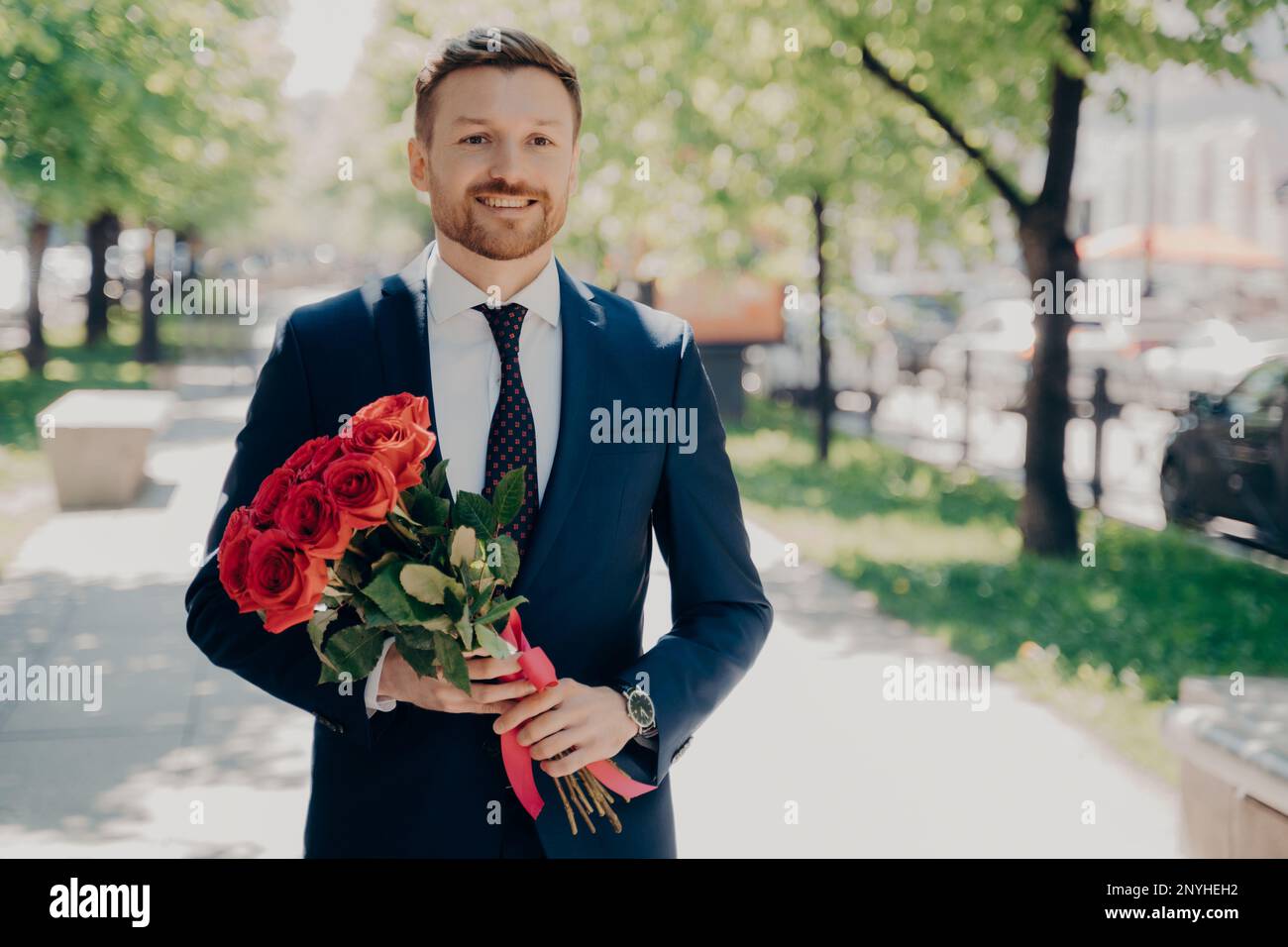 Ein lächelnder, stilvoller Gentleman im dunkelblauen Smoking mit einem großen Strauß roter Rosen, der an einem sonnigen Tag durch die grüne Gasse im Park spaziert, um seine Geliebte zu treffen Stockfoto