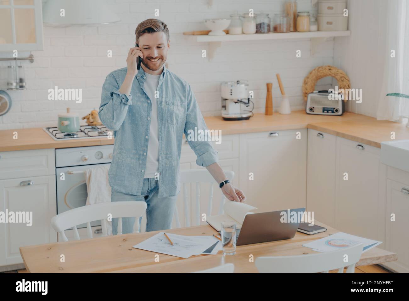Attraktiver, lächelnder, bärtiger Mann, der lässige Kleidung trägt und an einem Laptop arbeitet, während er zu Hause am Küchentisch steht und sich mit seinem Handy-Witz unterhält Stockfoto
