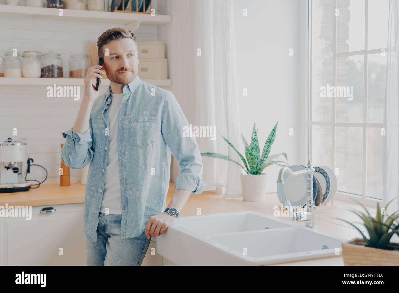 Junger Mann in lässigen Jeans Hemd, der mit seinem Partner am Handy spricht und wegschaut, während er an einem Holztisch in einer modernen Kücheneinrichtung steht Stockfoto