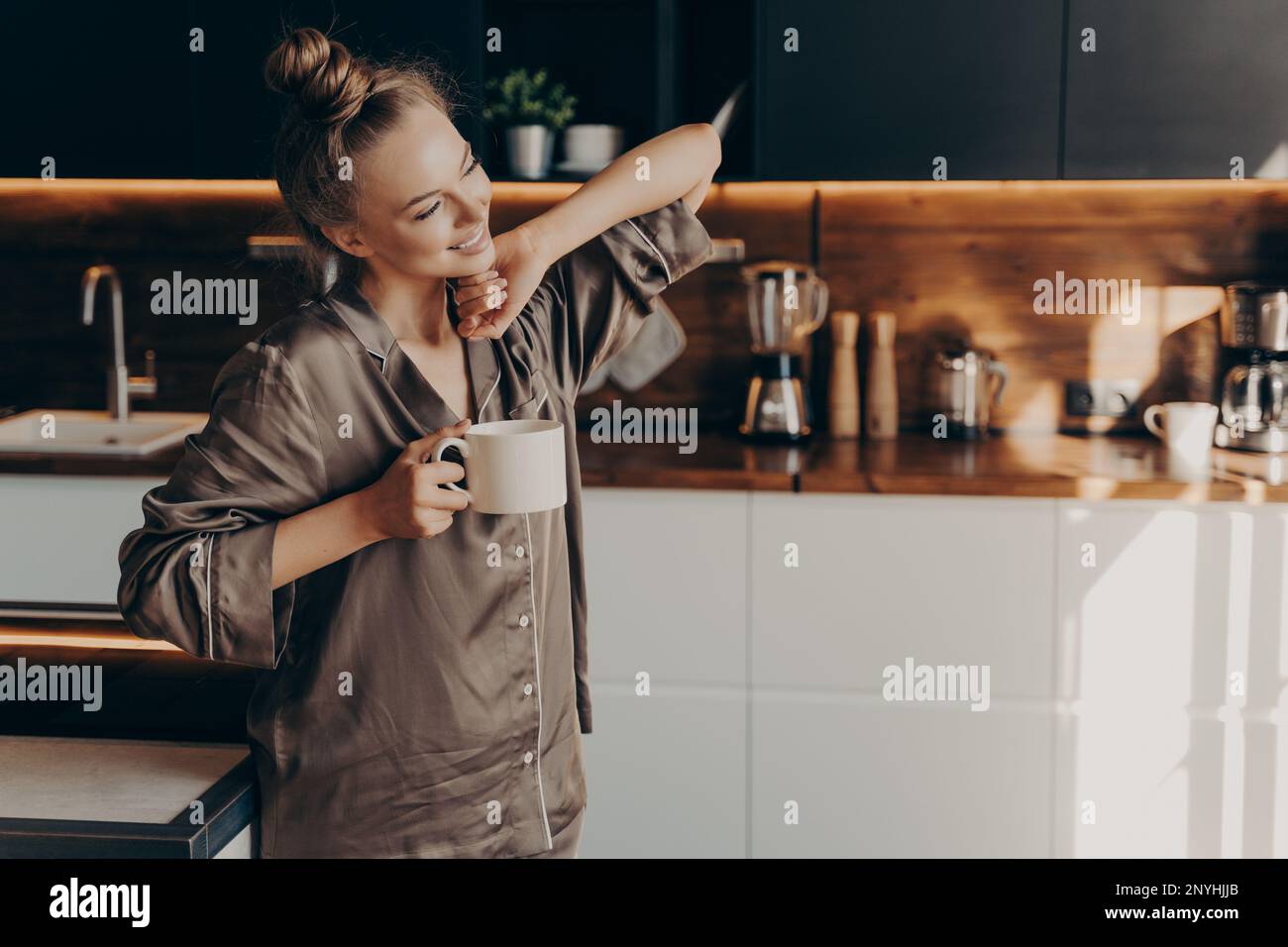 Eine junge, ziemlich entspannte Frau in einem bequemen Schlafanzug, die sich früh am Morgen aus dem Schlaf streckt, während sie eine Tasse Kaffee in der Hand hält und in einer stilvollen Küche steht Stockfoto