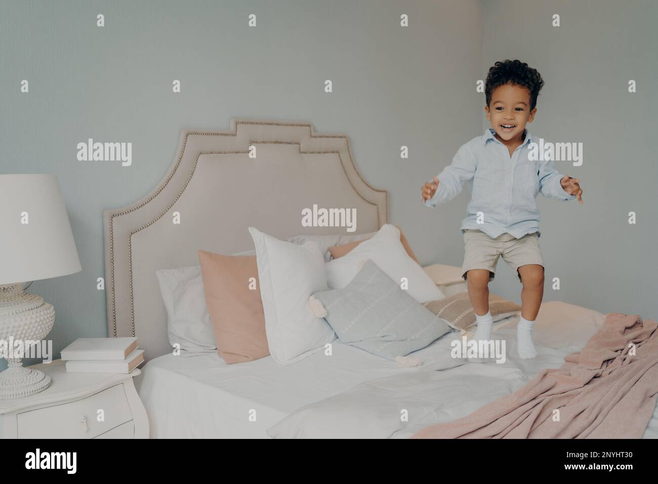 Ein Junge aus verschiedenen ethnischen Gruppen in lässiger Kleidung, der Spaß hat und auf ein großes Bett springt, mit weißen und pinkfarbenen Bettwäsche und Kopfkissen. In der Luft fotografiert Stockfoto