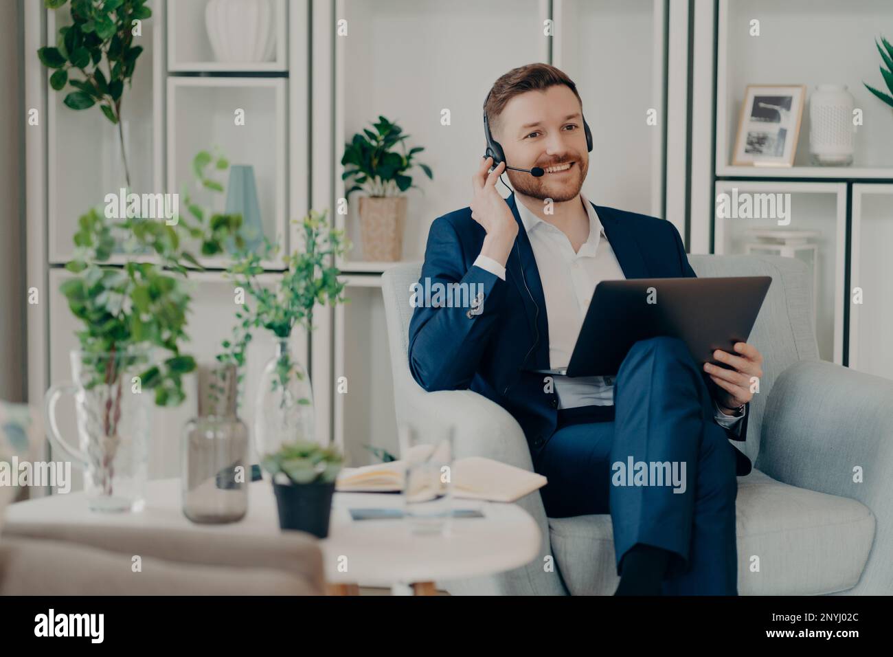 Freundlicher Unternehmensberater oder Vertriebsleiter im Anzug, der ein Notebook-Headset verwendet und mit dem Kunden im Wohnzimmer über Videokommunikation spricht Stockfoto