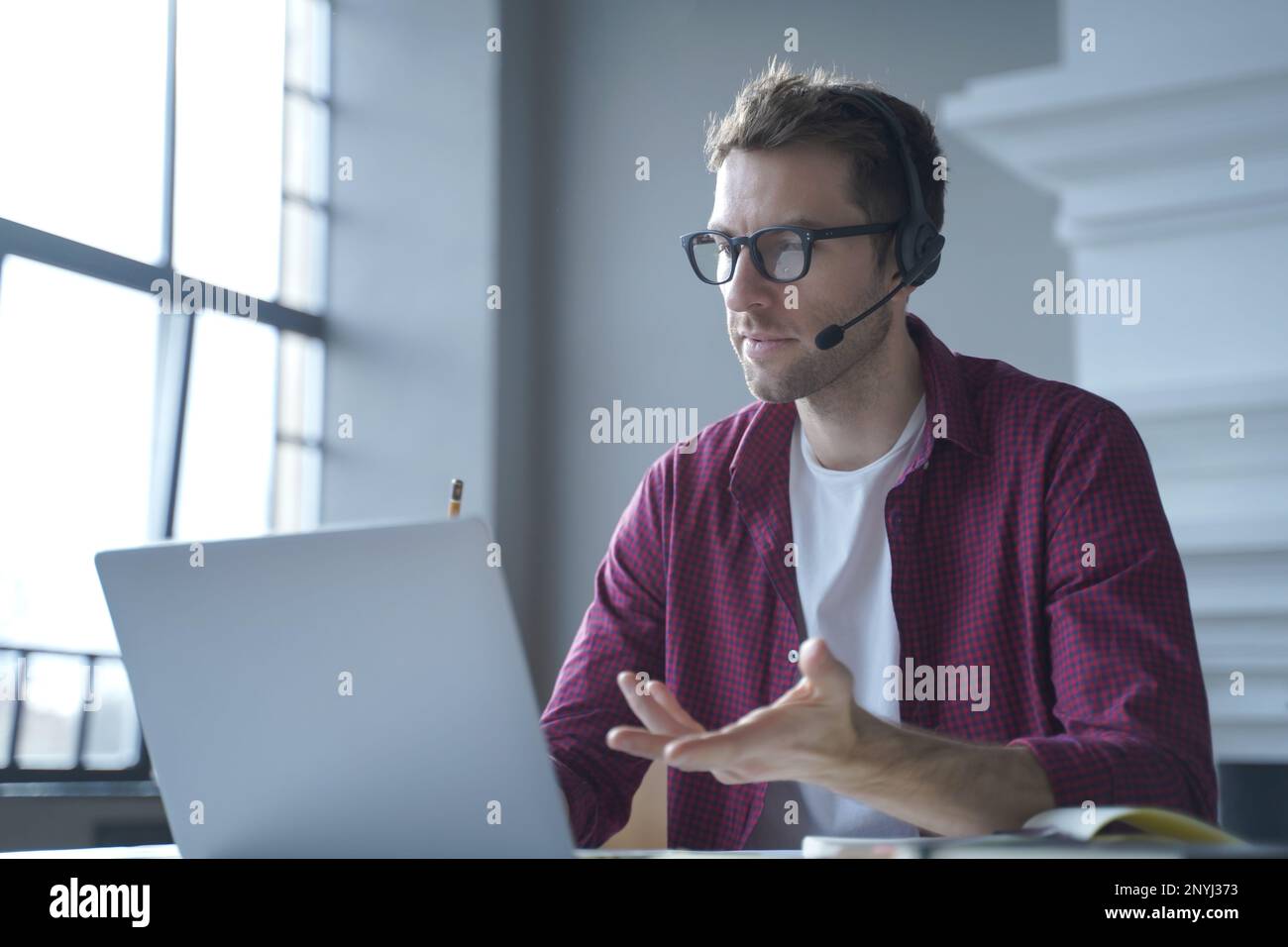 Europäischer Online-Lehrer mit Brille, sitzt am Schreibtisch in einem Headset und spricht per Videoanruf auf einem Laptop, während er von zu Hause aus arbeitet. Di Stockfoto