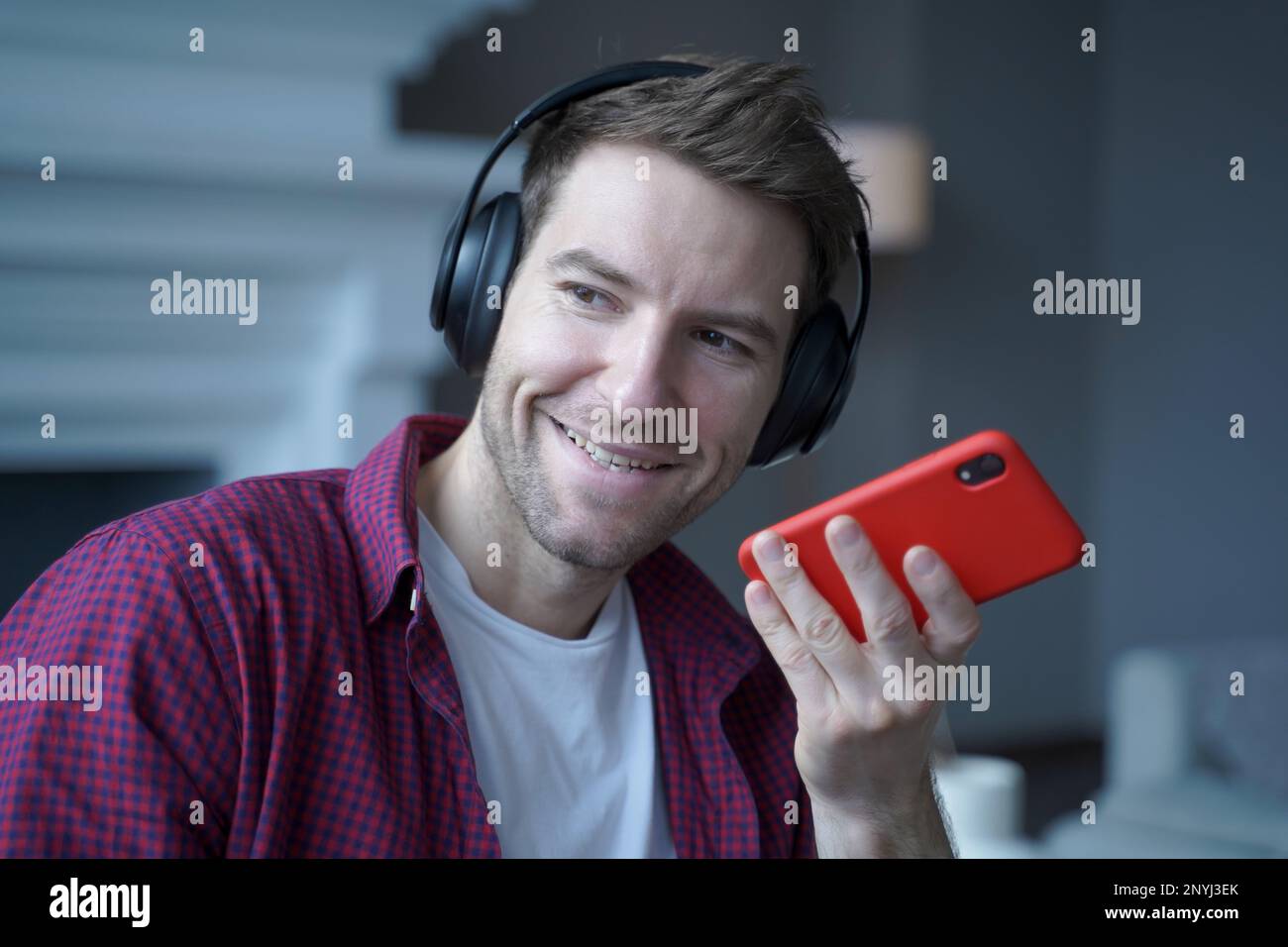 Lächelnder junger Mann, deutscher Sprachlehrer in Kopfhörern, der mit einem Schüler auf dem Handy spricht, fröhlicher österreichischer Typ, der eine Sprachnachricht für den Kollegen aufnimmt, uns Stockfoto