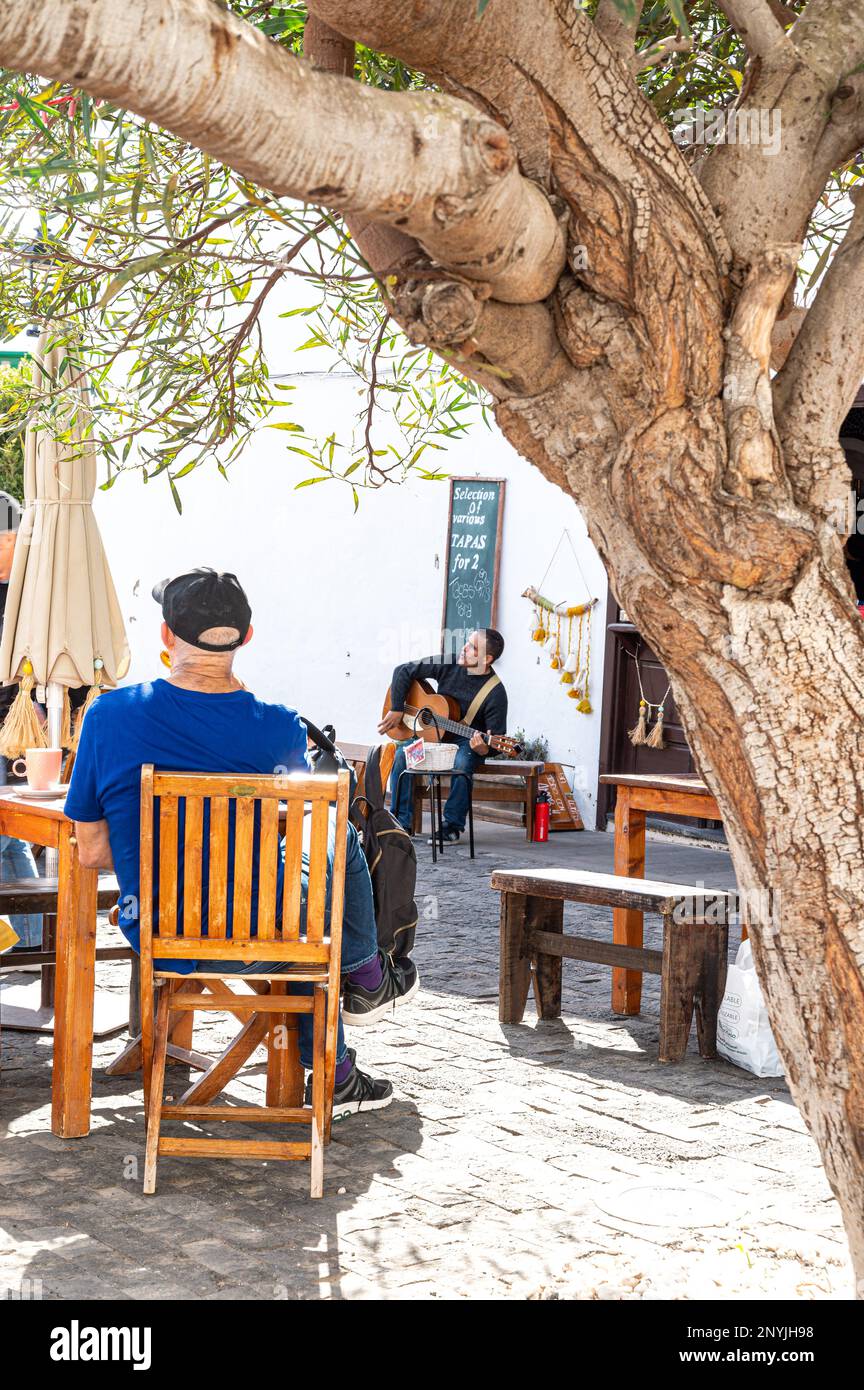 Perspektivenaufnahme eines Musikers, der in einem kleinen Café im Freien auftritt, mit einem Mann im blauen T-Shirt, der auf einem Stuhl am Baumstamm sitzt Stockfoto