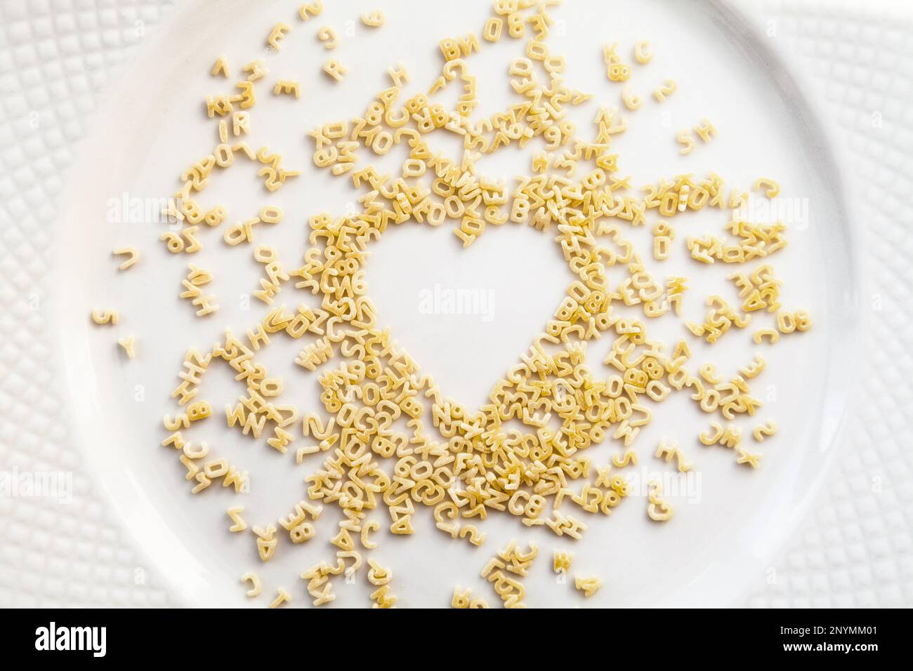 Chaotische Nudeln kleine Buchstaben und Ziffern in der Form eines Herzens auf einem weißen Teller verstreut. Ungekocht Trockenfutter Zutat Hintergrund. Liebe für cooki Stockfoto