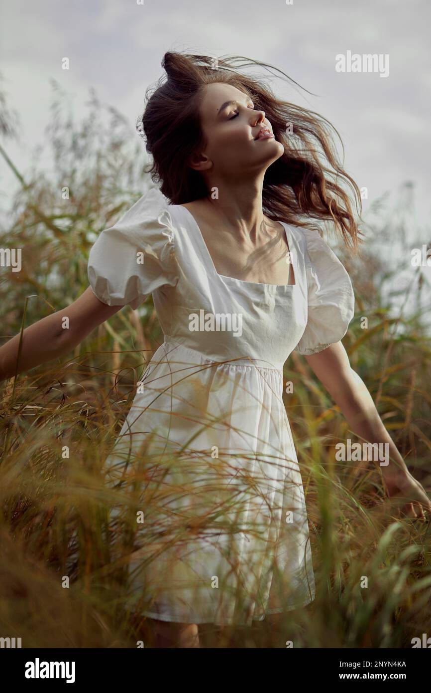 Romantische junge Frau auf dickem, hohem Gras auf dem Feld. Traumhafte, sanfte Frau in einem weißen Kleid in der Natur. Natürliche Schönheit, Freude am Leben Stockfoto