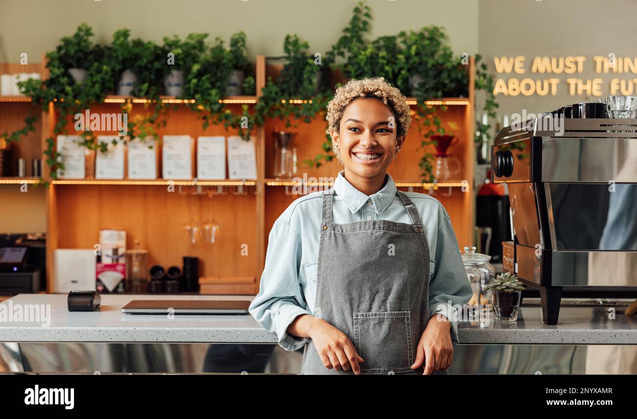 Selbstbewusster Besitzer eines Coffee Shops in einer Schürze. Junge lächelnde Frau in einer Schürze, die an der Bar steht. Stockfoto