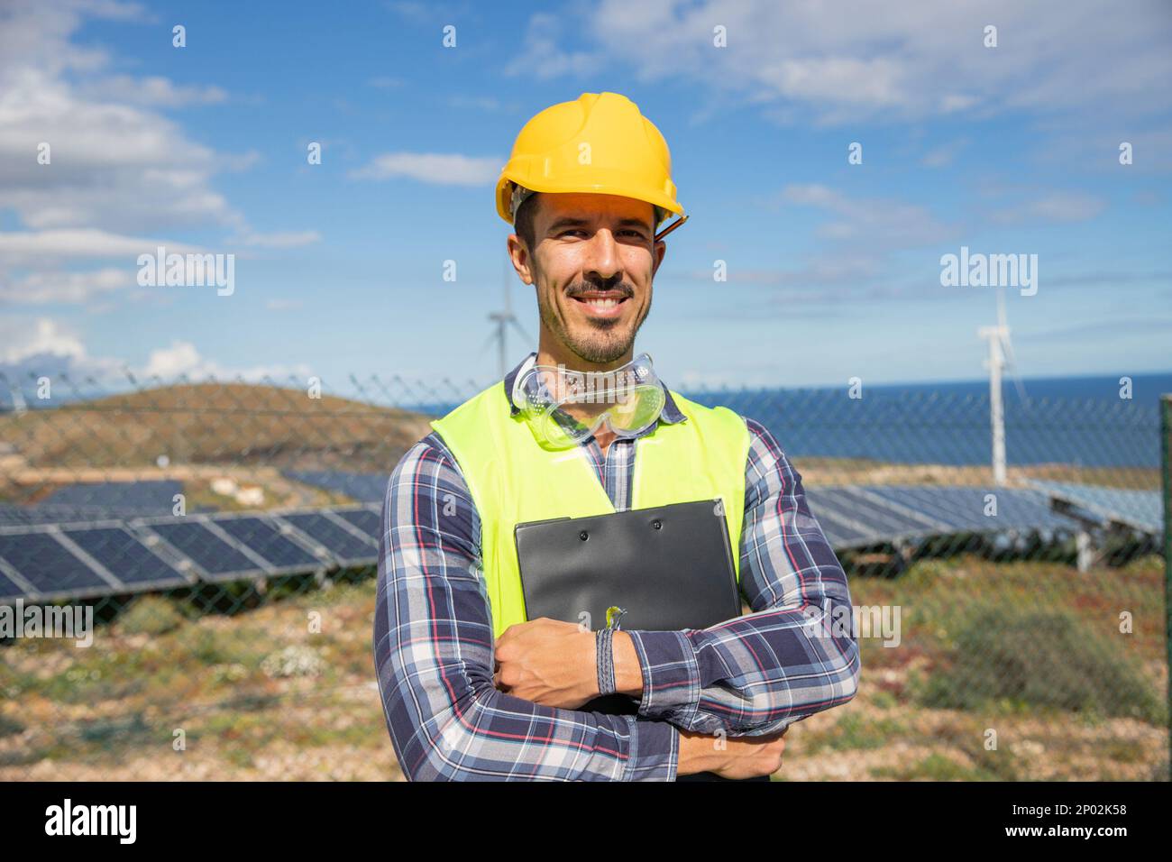 Ein lächelnder Ingenieur mit gefalteten Armen auf einer Solarfarm, der einen Ordner in der Hand hält. Stockfoto