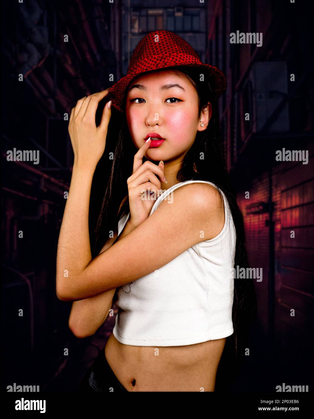 Teenager-Mädchen mit roter Fedora, die in der Gasse steht, mit Händen auf den Lippen und Hut | Rote Töne | Moody Stockfoto