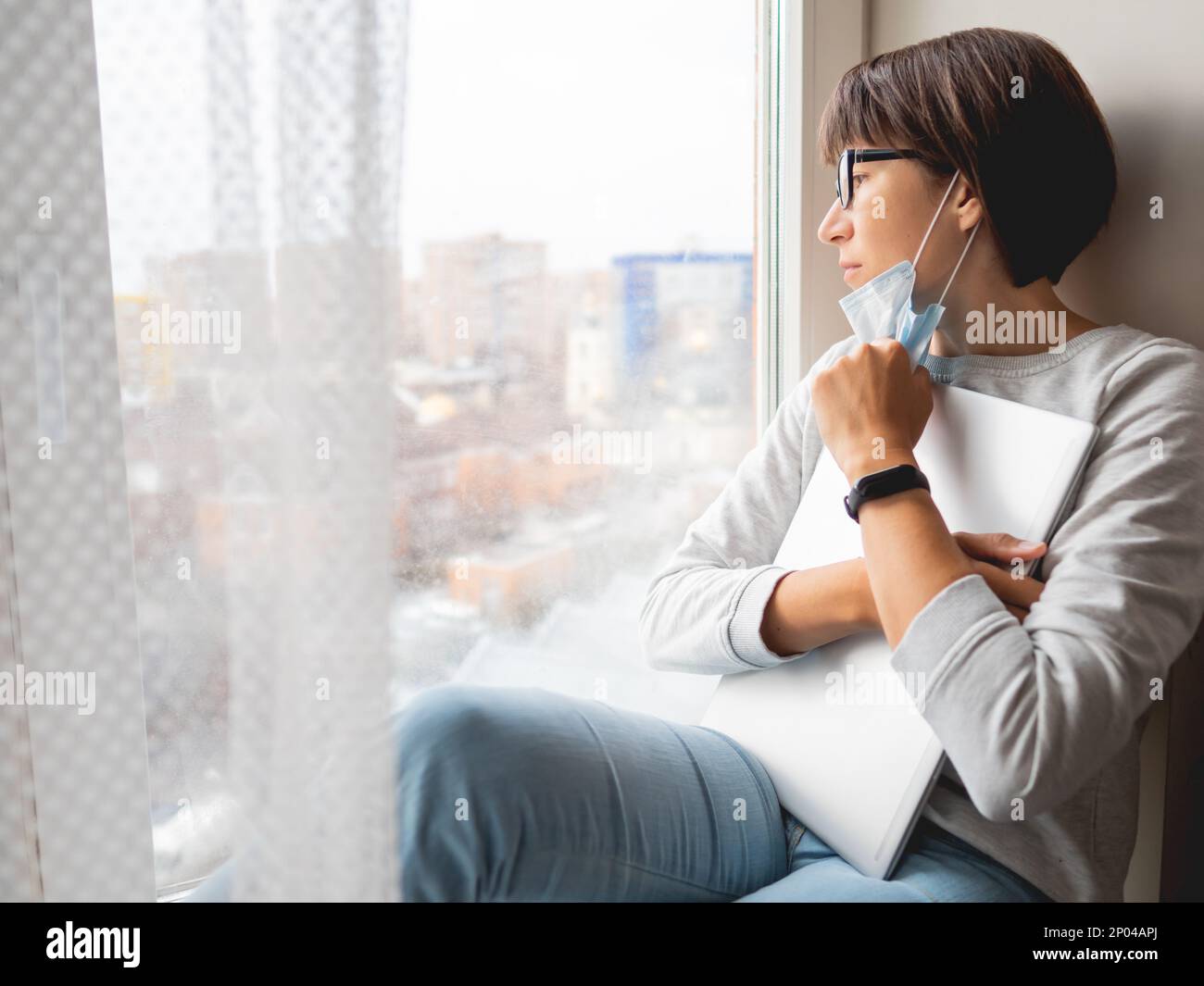 Eine Frau mit medizinischer Maske hatte ihren Job verloren. Sie sitzt auf der Fensterbank mit geschlossenem Laptop. Sperrung Quarantäne wegen Coronavirus COVID19. Selbstisolation Stockfoto