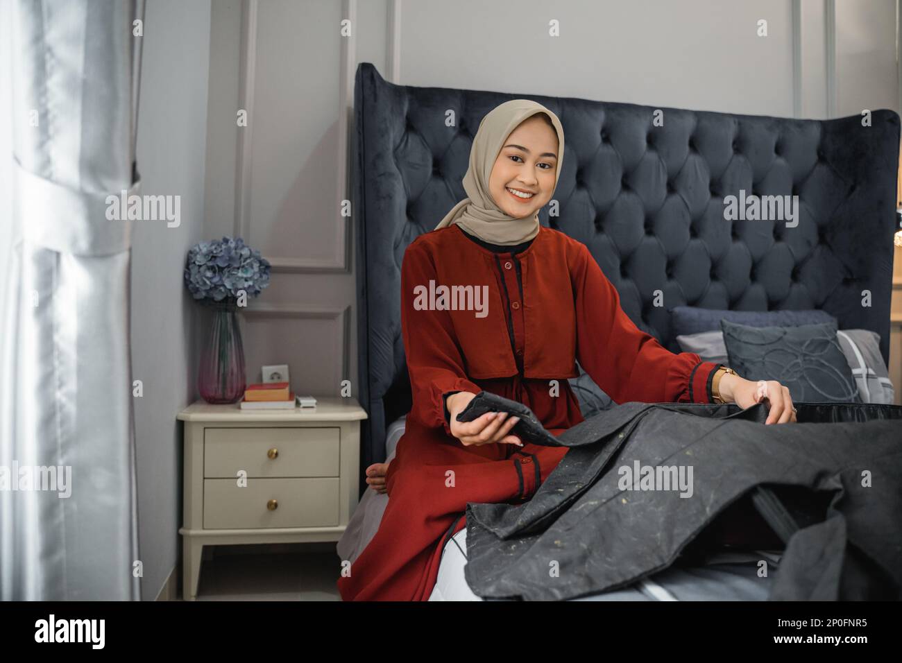 Eine Frau im Hijab lächelt, während sie Kleider zusammenfaltet und auf dem Bett sitzt Stockfoto