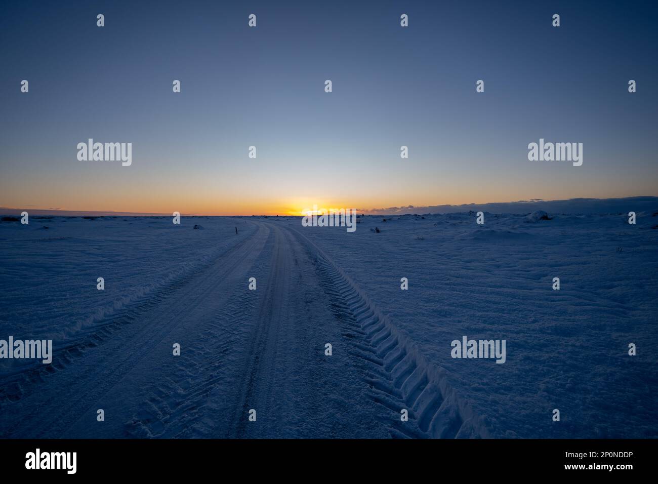 Straße und flache verschneite Landschaft in fast Dunkelheit mit Reifenspuren, die zum Horizont führen, während die Sonne in einem magischen goldenen isländischen Sonnenaufgang aufgeht. Stockfoto