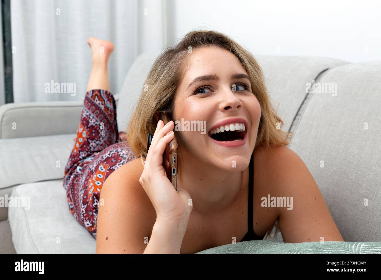 Die junge blonde Frau führt ein freundliches Gespräch am Telefon, während sie sich auf der Couch mit einem Lächeln im Gesicht ruht. Stockfoto