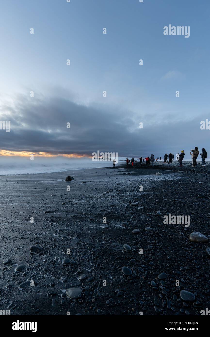 Reynisfjara schwarzer Sandstrand bei Sonnenuntergang beleuchtet von den letzten Sonnenstrahlen vom Horizont des Meeres mit Touristen, die begeistert sind und Fotos machen Stockfoto