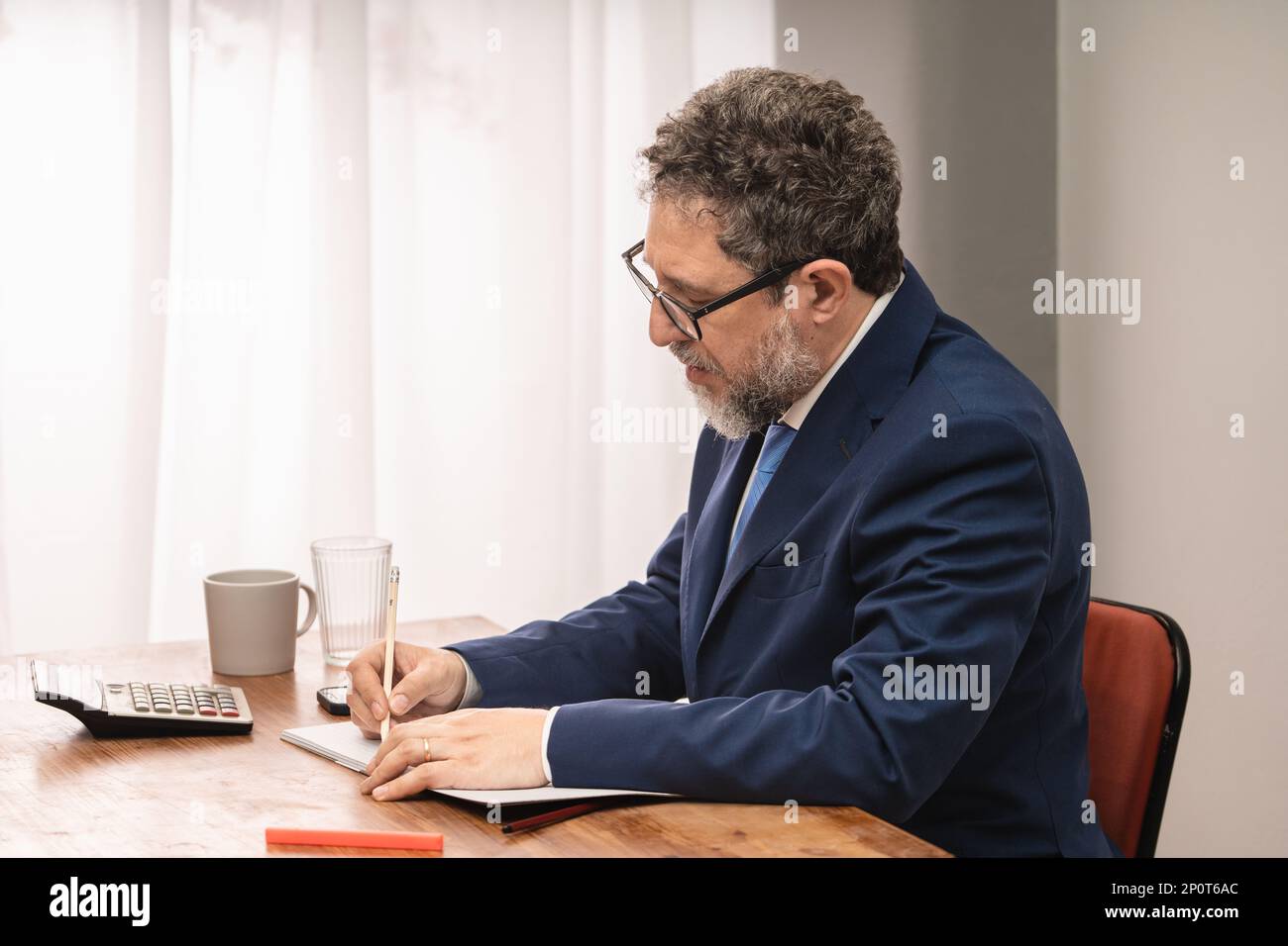 Geschäftsmann in den 50s Jahren mit Bart, Anzug und Krawatte, sitzt am Schreibtisch und schreibt mit einem Bleistift auf einem Notizblock Stockfoto