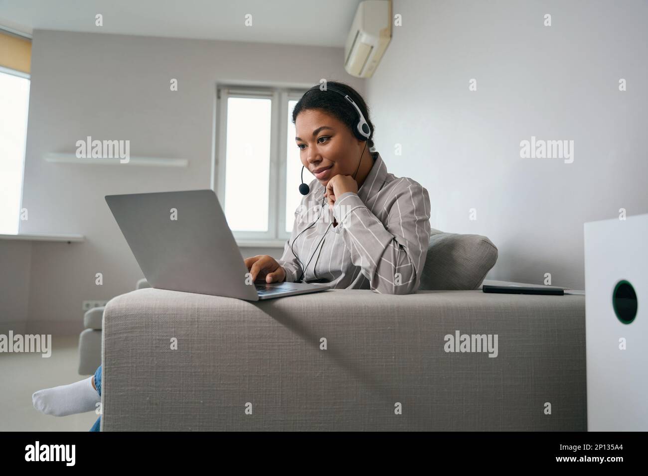Eine Frau mit Headset auf dem Kopf sitzt auf dem Sofa mit einem Laptop Stockfoto