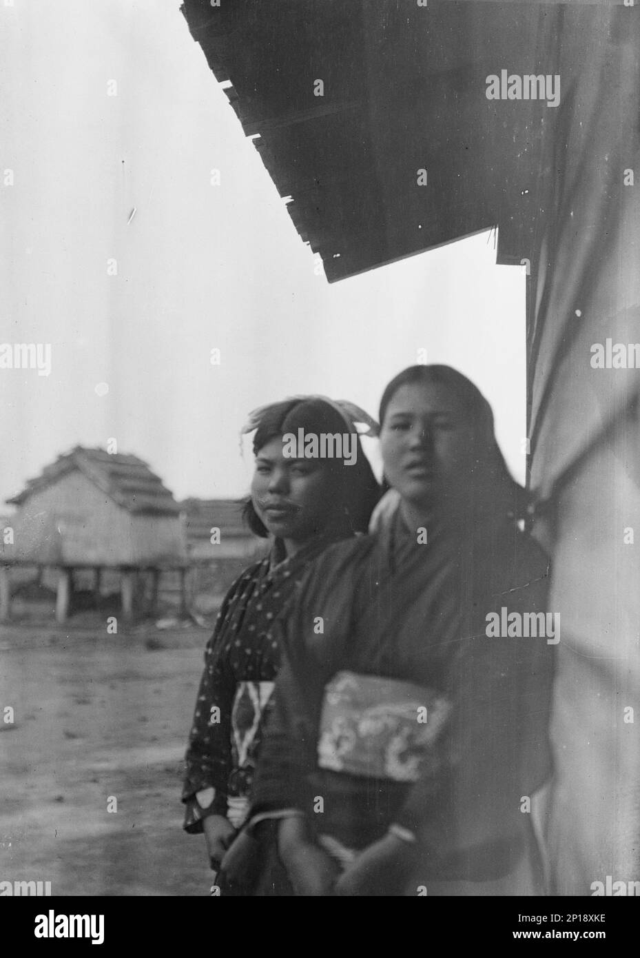 Zwei Ainu-Frauen stehen draußen an der Wand einer Holzhütte, 1908. Stockfoto
