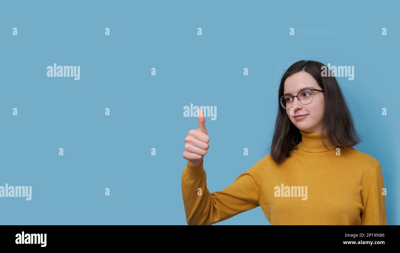 Ein Porträt einer charmanten, ernsthaften Schülerin mit Brille und gelbem Pullover, die ihren Finger auf blauem Hintergrund hebt. Eine ähnliche Geste. Das Mädchen Stockfoto