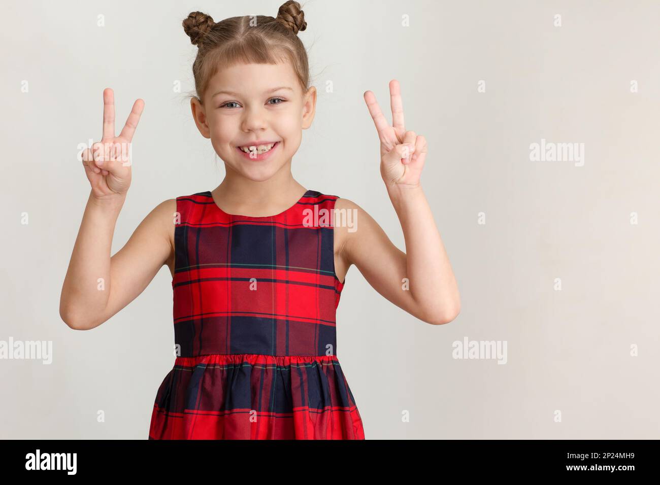 Lächelndes Kind kleines Mädchen mit dem Schild V Victory auf beiden Händen, weiß-weißes Kind von 6 7 Jahren in rotem Karomuster auf grauem Hintergrund Stockfoto