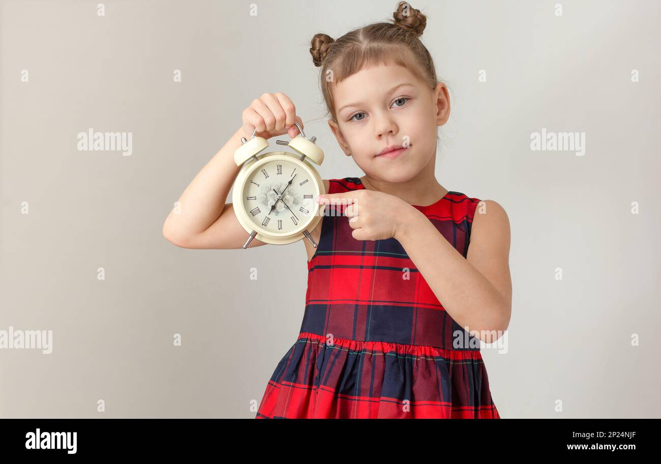 Ernsthaftes Kind, das den Wecker hält und Zeigefinger zeigt, Konzept, früh aufzuwachen, weiß-kleines Mädchen von 6 7 Jahren in rotem Karomuster Stockfoto