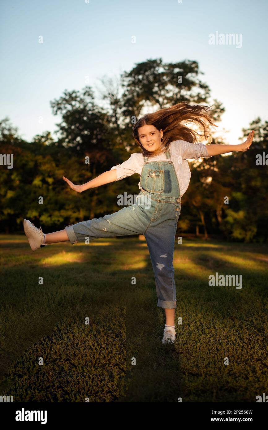 Ein Schulmädchen in Jeansanzug springt und hat Spaß auf dem Rasen mit grünem Baumhintergrund bei Sonnenuntergang Stockfoto