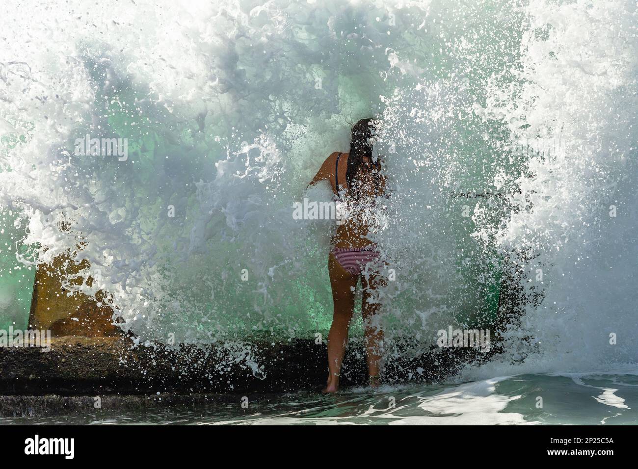 Ein Mädchen, das sich nicht wiedererkennt, steht am Strand am Gezeitenschwimmbad, steht Kopf an der Wand und begegnet einem Hauch von Meereswellen, die von Wildwasser explodieren. Stockfoto