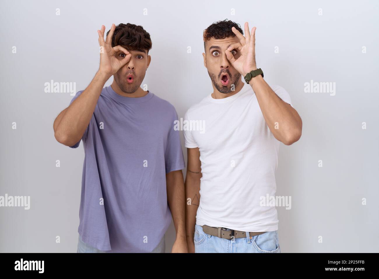 Homosexuelles schwules Paar, das über weißem Hintergrund steht und eine gute Geste macht, schockiert mit einem überraschten Gesicht, Augen, die durch die Finger schauen. Unglaublicher Ausdruck Stockfoto