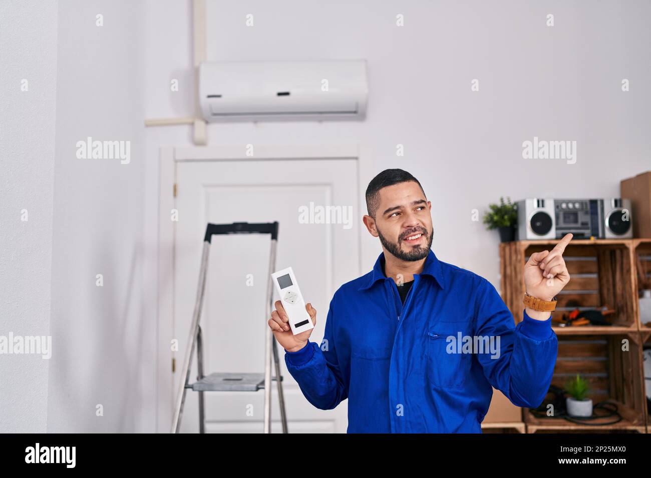 Hispanischer Servicetechniker, der mit der Klimaanlage arbeitet und lächelt, zeigt mit der Hand und dem Finger zur Seite Stockfoto