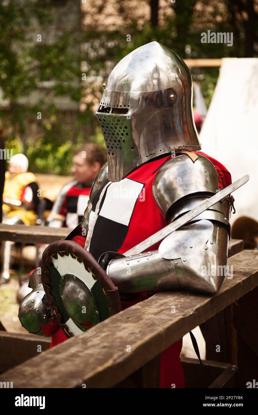 Kostüm aus der Mittelalterzeit beim Ritterturnier. Mittelalterliche historische Nachstellung - ein Mann mit Metallhelm und roter Rüstung, mit einem Schwert und Stockfoto