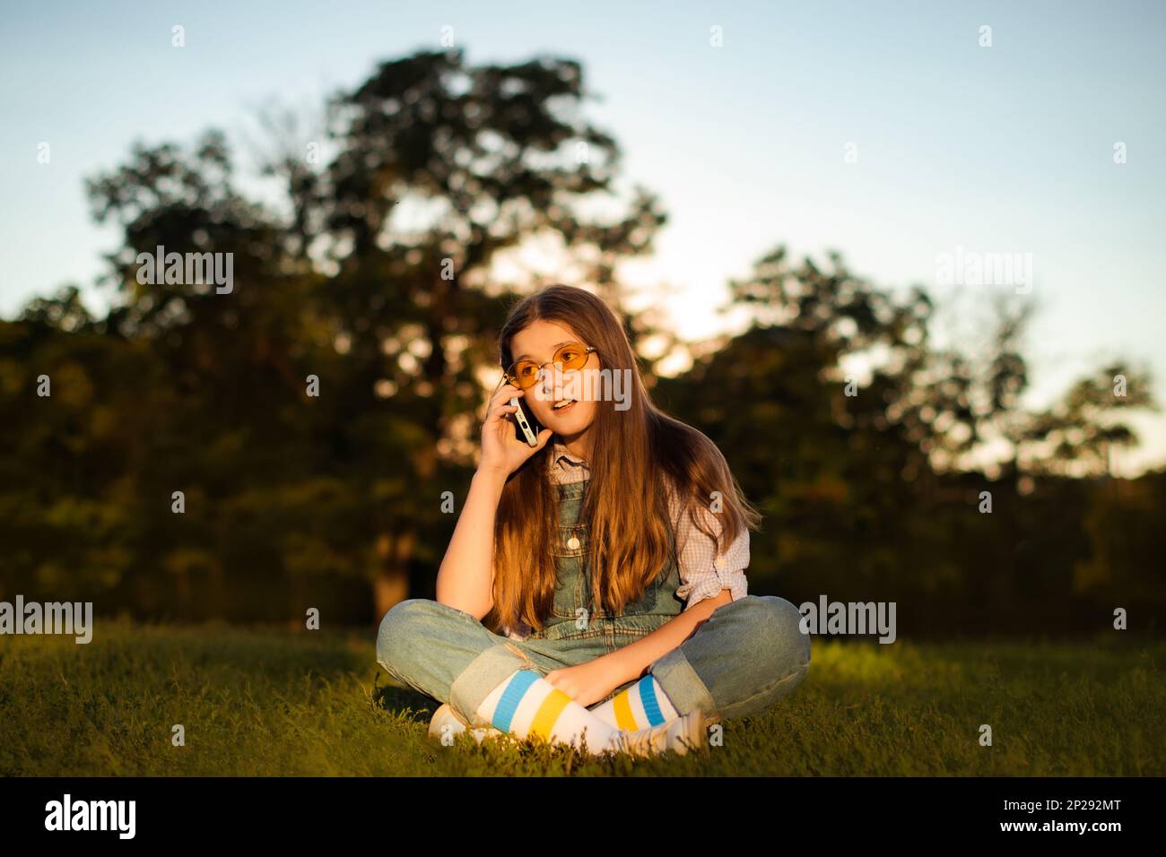 Schulmädchen in Denim-Overalls spricht auf Smartphone und sitzt auf grünem Gras im Park Stockfoto