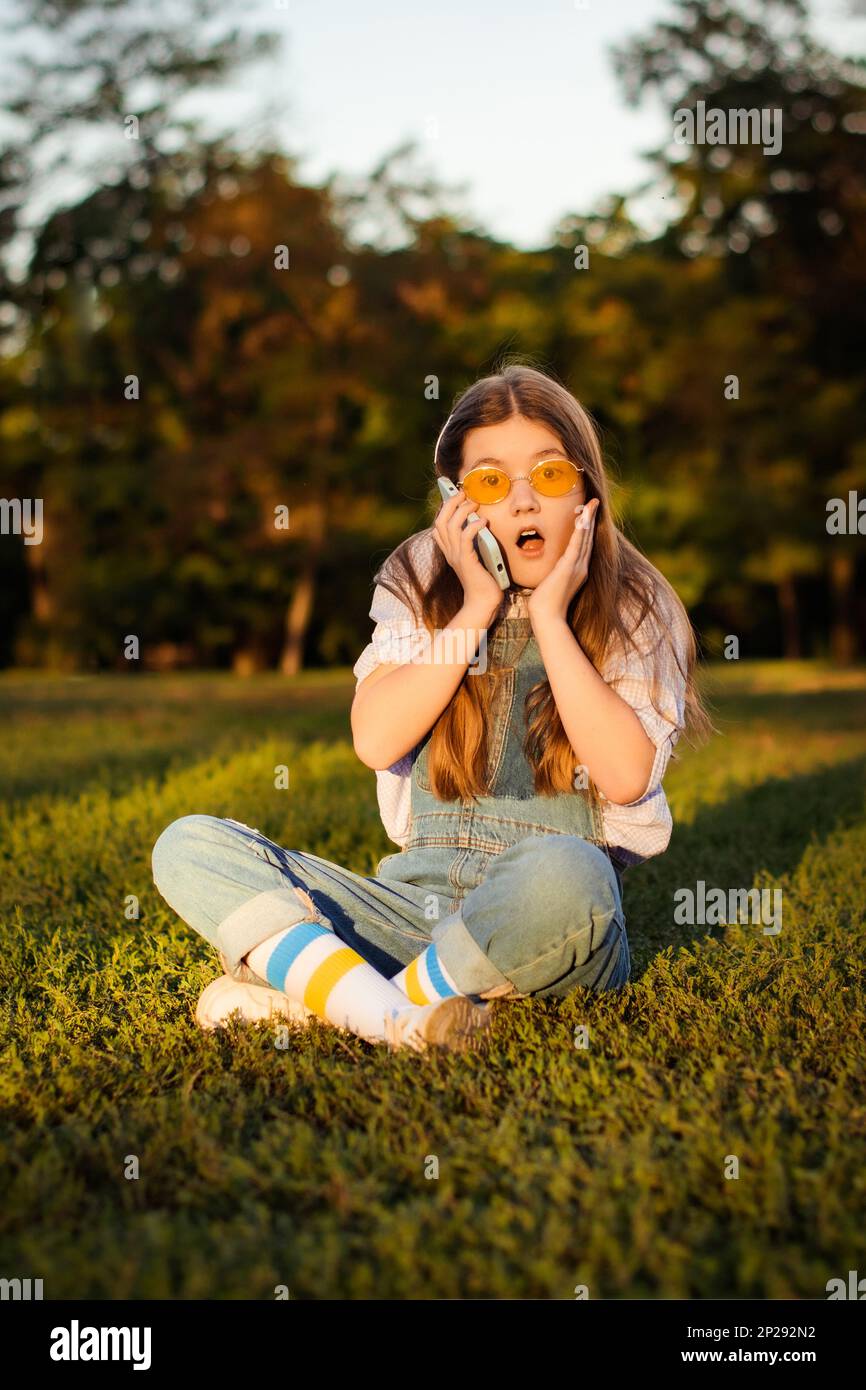 Überraschtes und aufgeregtes Schulmädchen in Denim-Overalls spricht auf einem Smartphone, das auf grünem Gras im Park sitzt Stockfoto