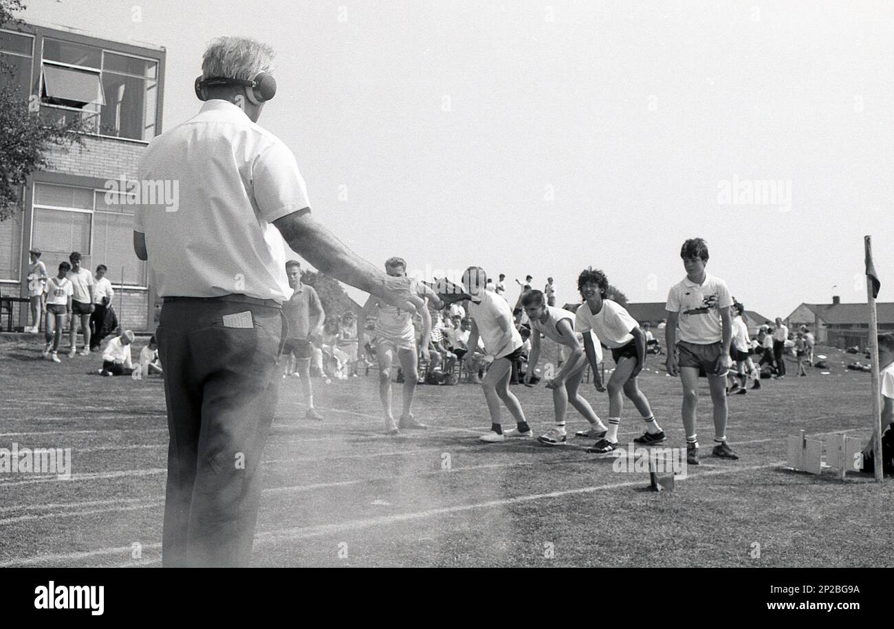 1989, Schulsporttag, draußen auf einem Sportplatz, männliche Schüler der Sekundarstufe bereit für ein Rennen, als männlicher Lehrer mit Gehörschutz, der Starter, wenn Rauch erscheint, feuert die Startpistole, England, Großbritannien. Stockfoto
