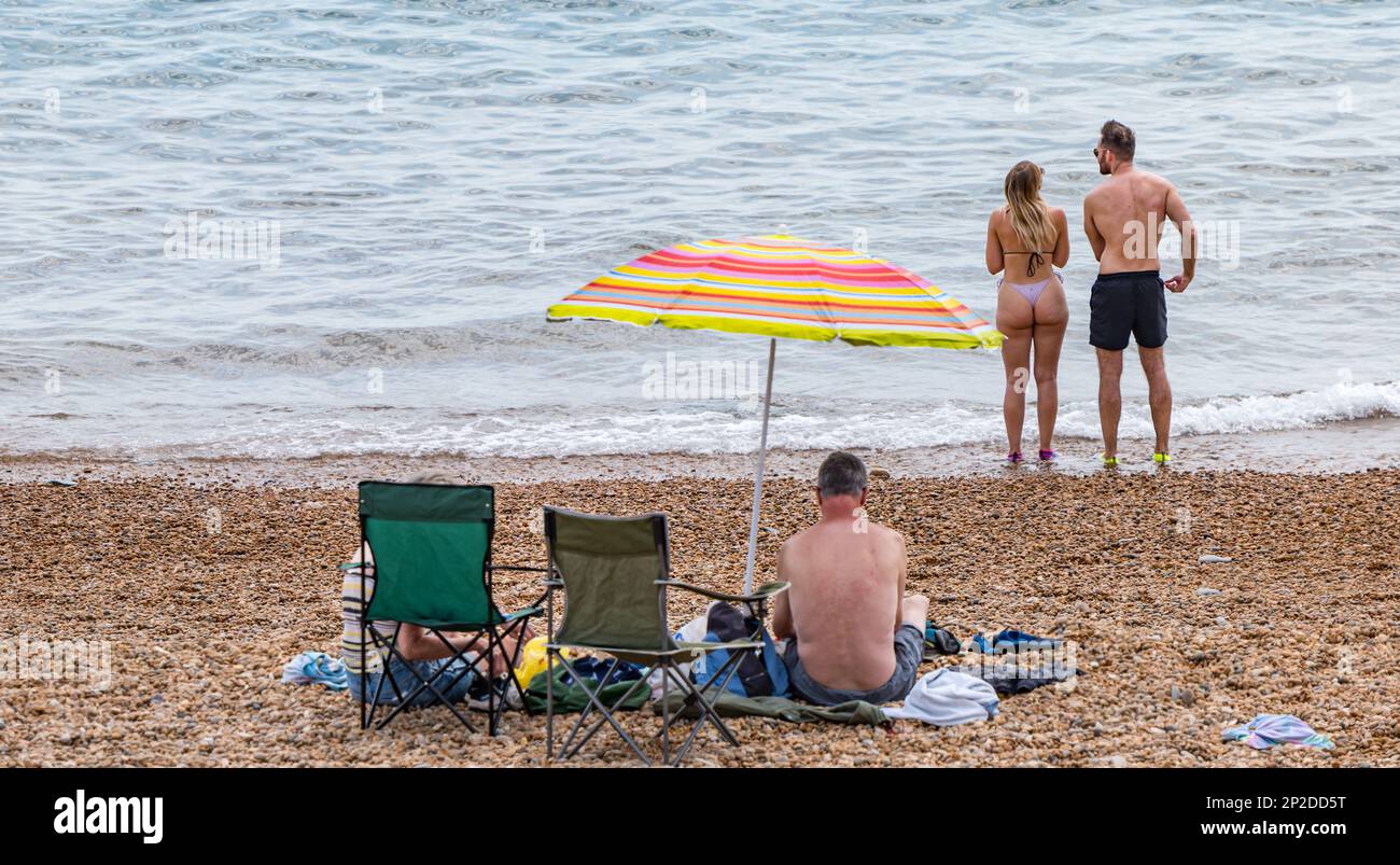 Ein Paar am Strand mit einer jungen Frau, die einen Bikini-Badeanzug am Seatown Beach in Summer an der Jurassic Coast, Dorset, England, UK trägt Stockfoto
