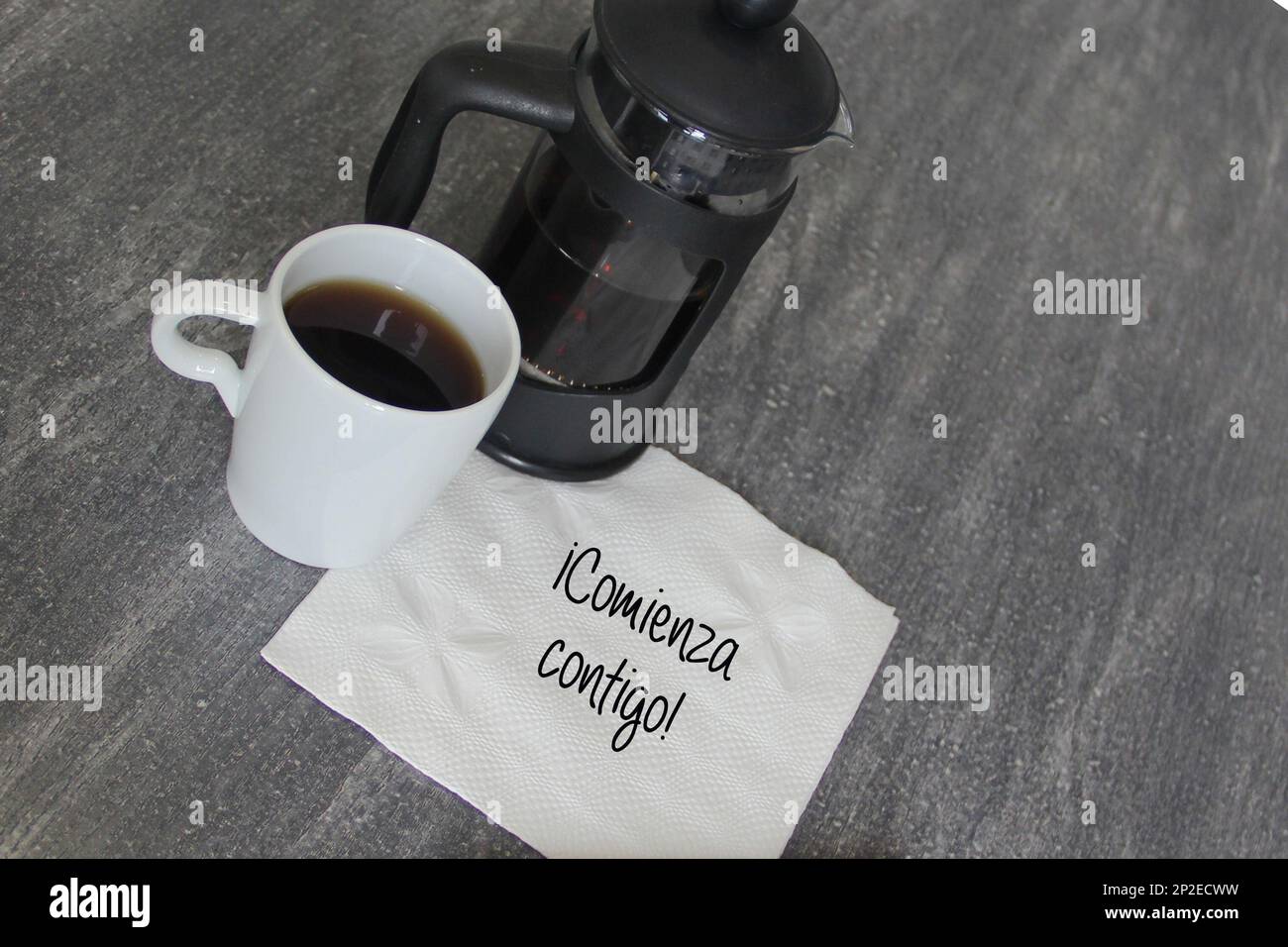 Eine Tasse Kaffee, eine französische Presse und eine weiße Serviette mit der Nachricht „IT fängt mit dir an!“ in spanischer Sprache Stockfoto