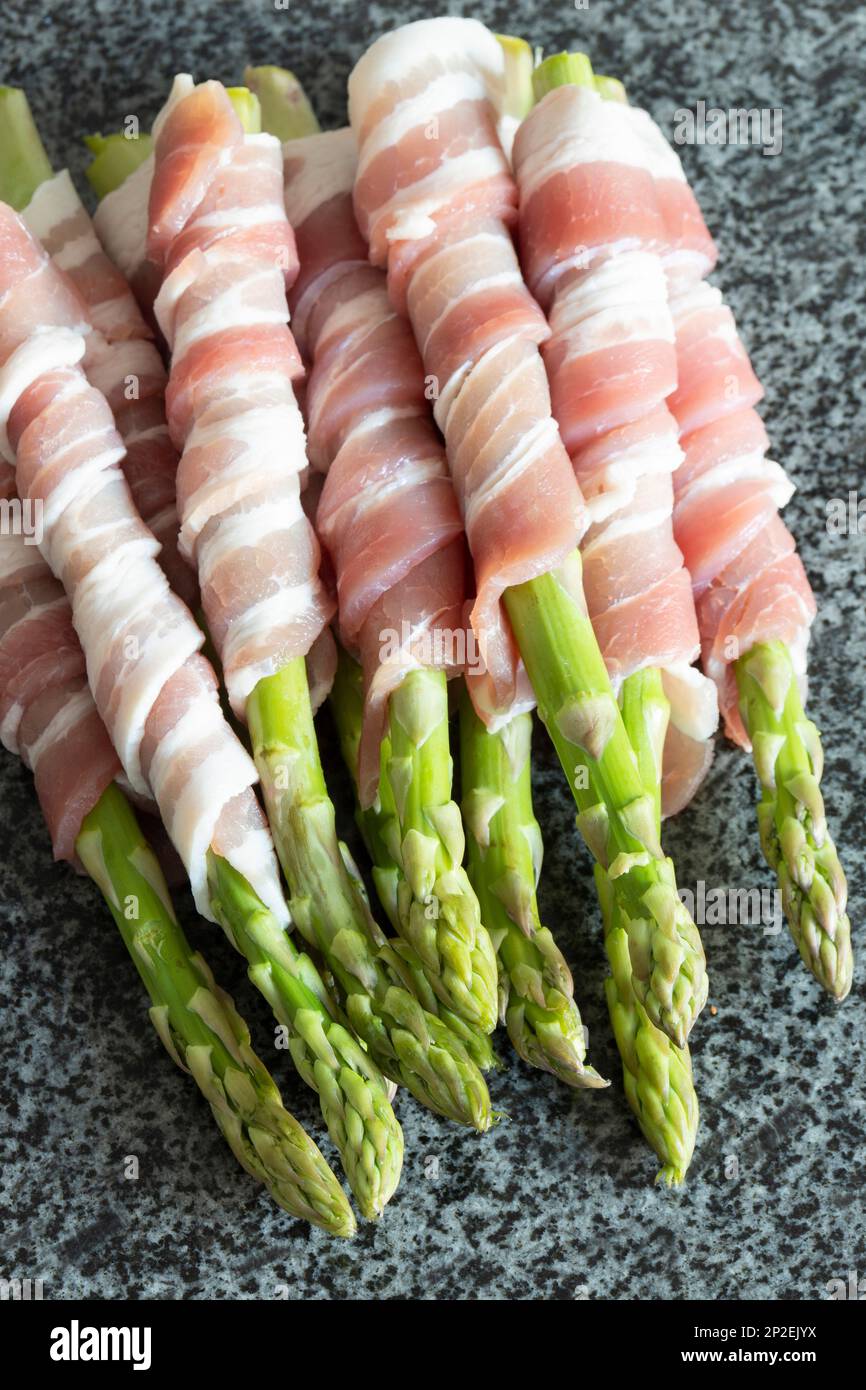 Roher organischer Spargel, in durchwachsenem Speck verpackt, auf einer schwarzen Granitarbeitsfläche. Konzept mit frischen saisonalen Produkten Stockfoto