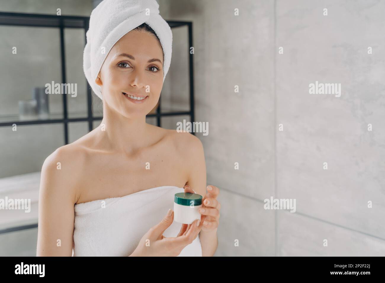 Hübsche hispanische Frau, die ein Modell-Kosmetikglas mit Sahne oder Serum hält, Werbung für Hautpflege Feuchtigkeits-Kosmetikprodukt. Frau im Handtuch nach dem Duschen Stockfoto