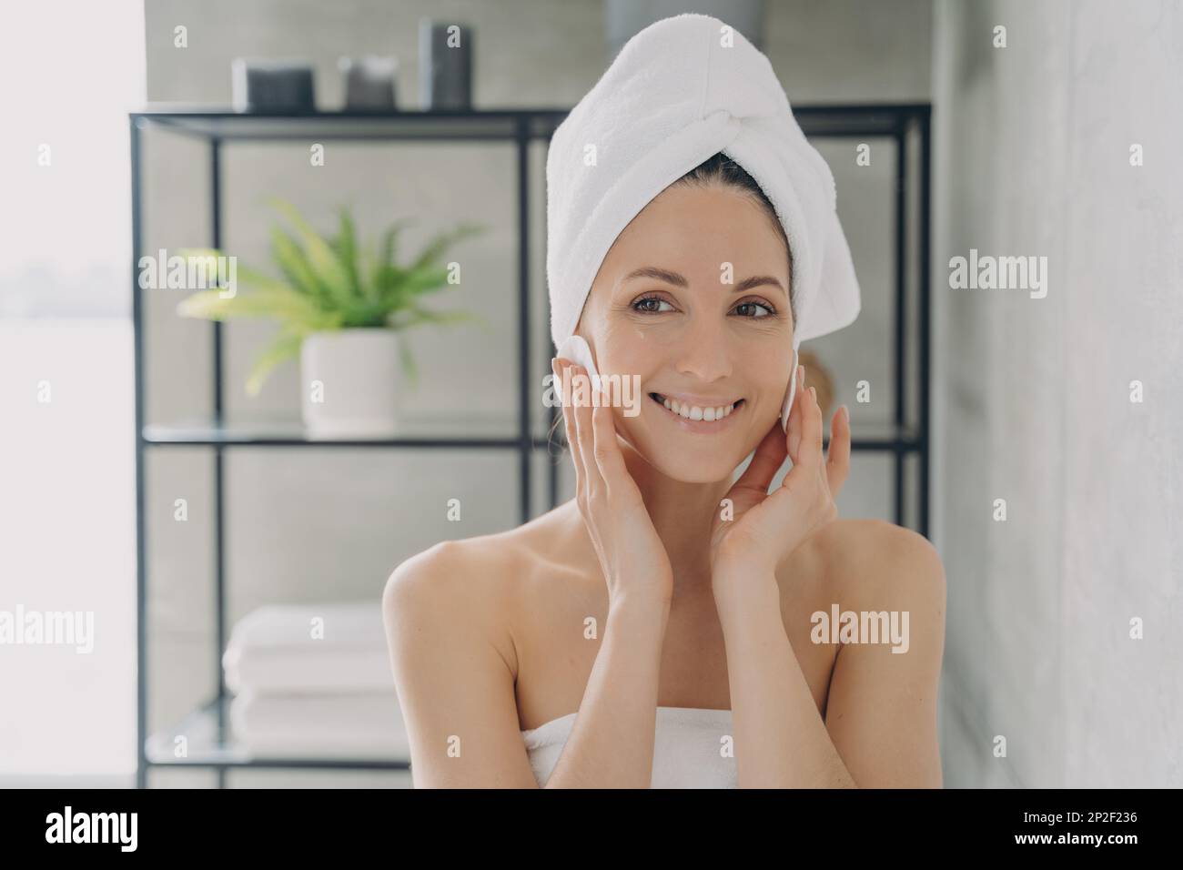 Frau mit Handtuch auf dem Kopf reinigt die Gesichtshaut und entfernt Make-up nach dem Duschen mit Wattepads. Latina-Frau genießt frische, saubere, gesunde Haut. Hautpflege b Stockfoto