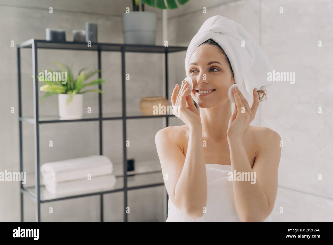 Hübsches lateinamerikanisches Mädchen, das die Gesichtshaut mit Wattepads reinigt. Eine lächelnde Frau, die in Handtücher eingewickelt ist, wischte das Make-up nach der Dusche im Badezimmer ab. Hautpflege da Stockfoto