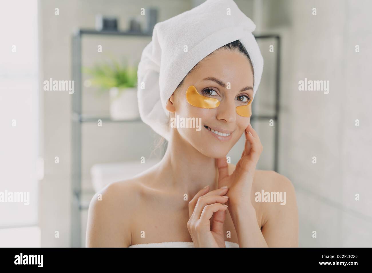Lächelnde hübsche hispanische Frau, die ein Handtuch trägt und goldene feuchtigkeitsspendende Hydrogelflecken auf der Unterhaut im Badezimmer trägt. Frau genießt Morgenantiagin Stockfoto