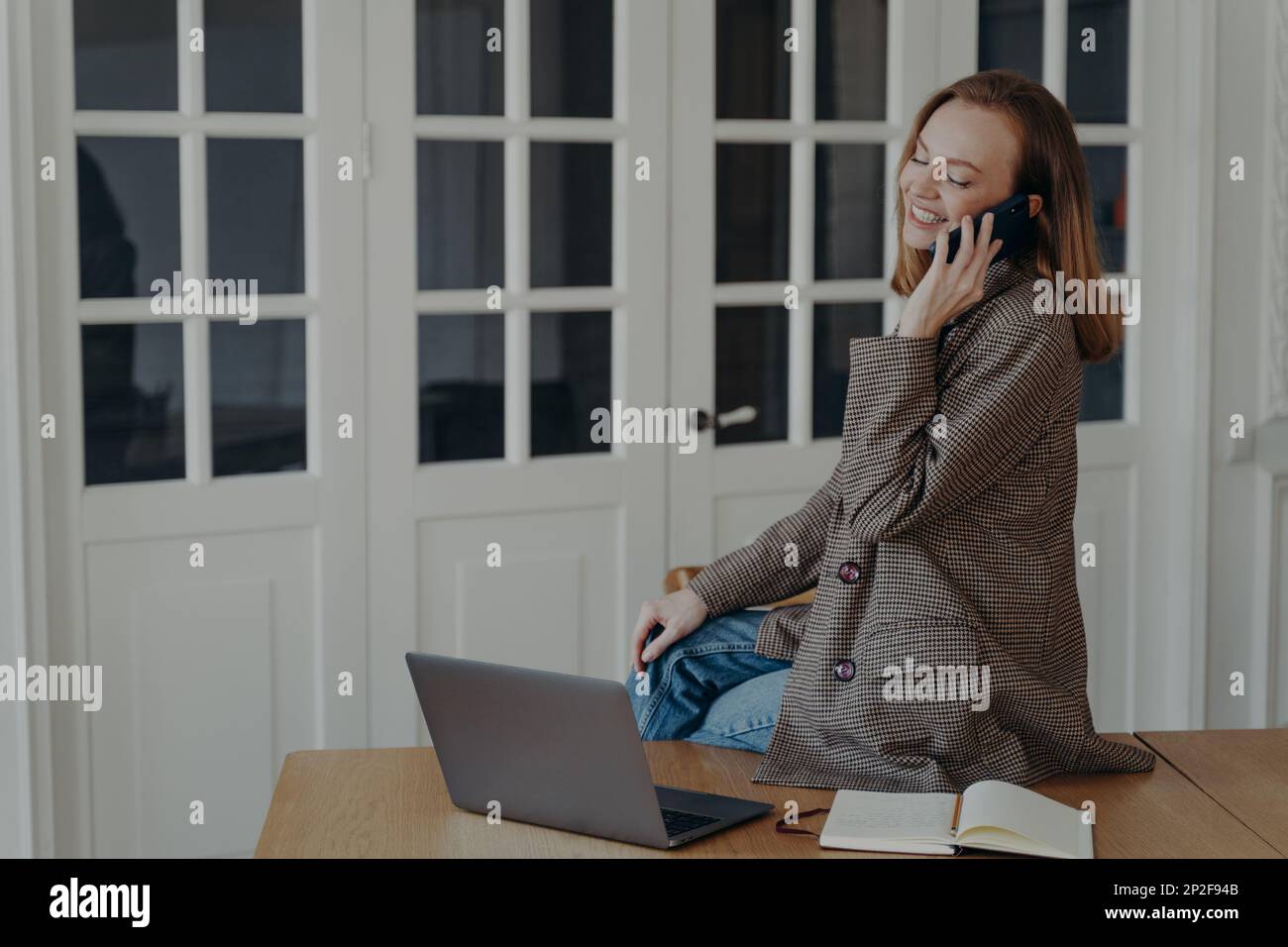 Eine lächelnde Freiberuflerin der Managerin beantwortet Anrufe auf dem Handy und sitzt mit einem Laptop am Telefon auf dem Schreibtisch. Glückliche junge Mitarbeiterin sitzt am Bürotisch Cha Stockfoto