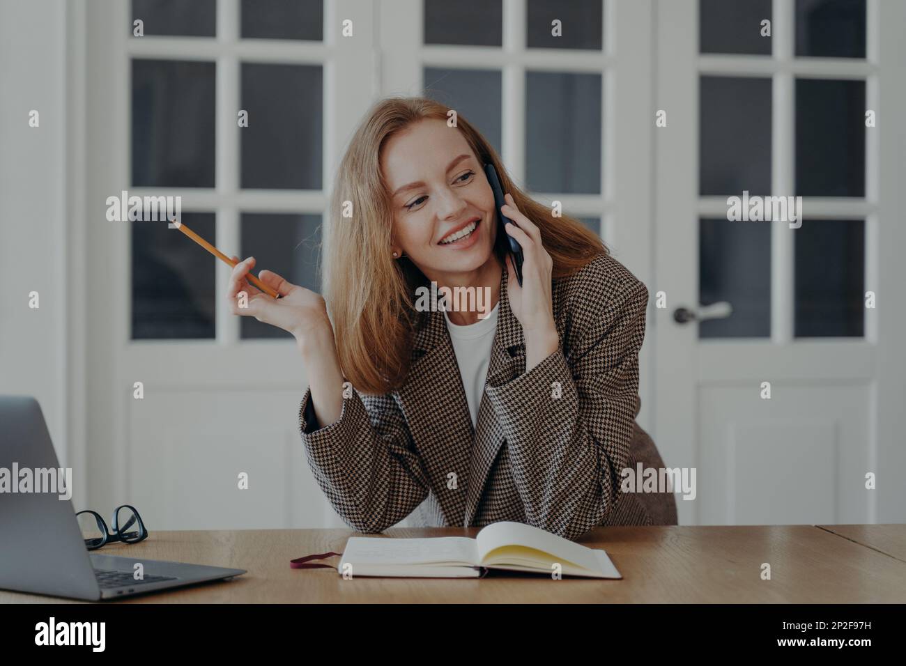 Lächelnde junge Geschäftsfrau spricht am Telefon, schreibt Notizen, sitzt am Schreibtisch. Weibliche Sekretärin mit Smartphone Stockfoto