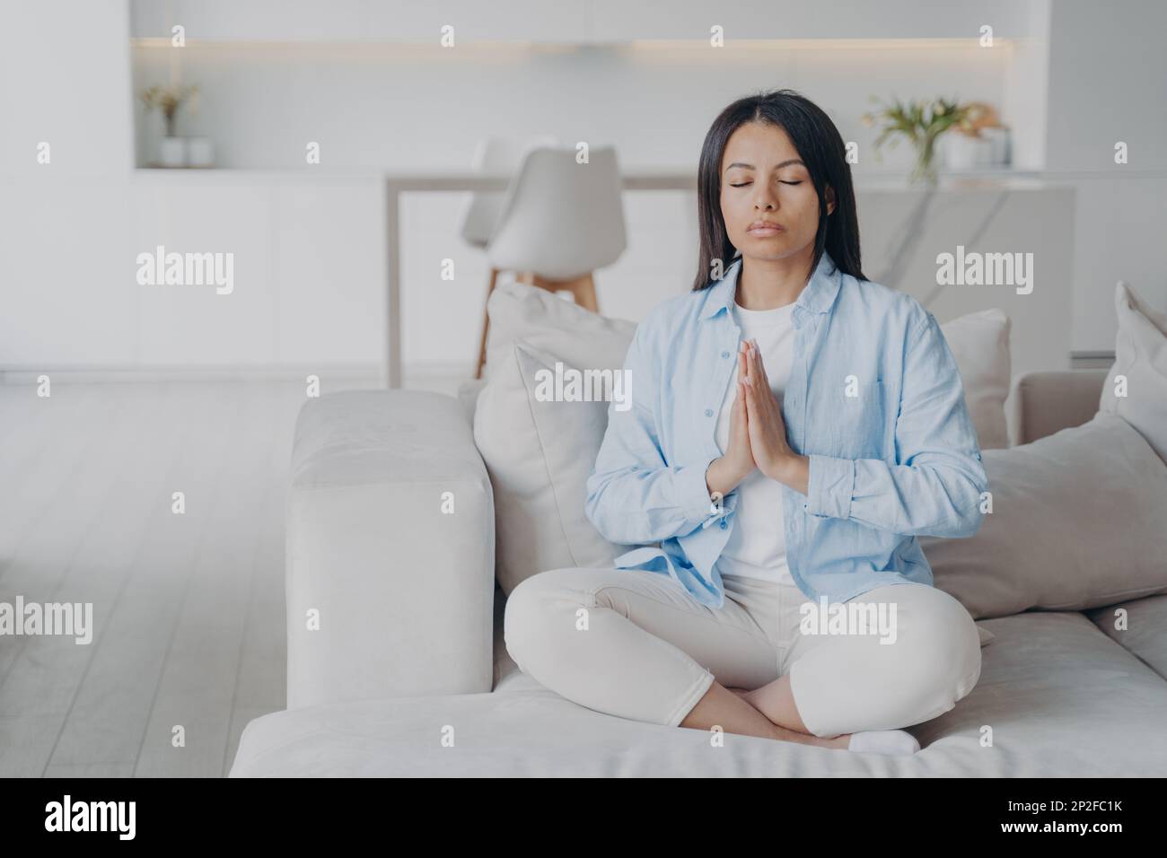 Ruhige Frau übt Yoga, meditiert, sitzt in Lotus-Pose zu Hause auf dem Sofa. Die Frau sitzt auf der Couch und entspannt die gefalteten Hände in einer namensfreundlichen Geste. Gut Stockfoto