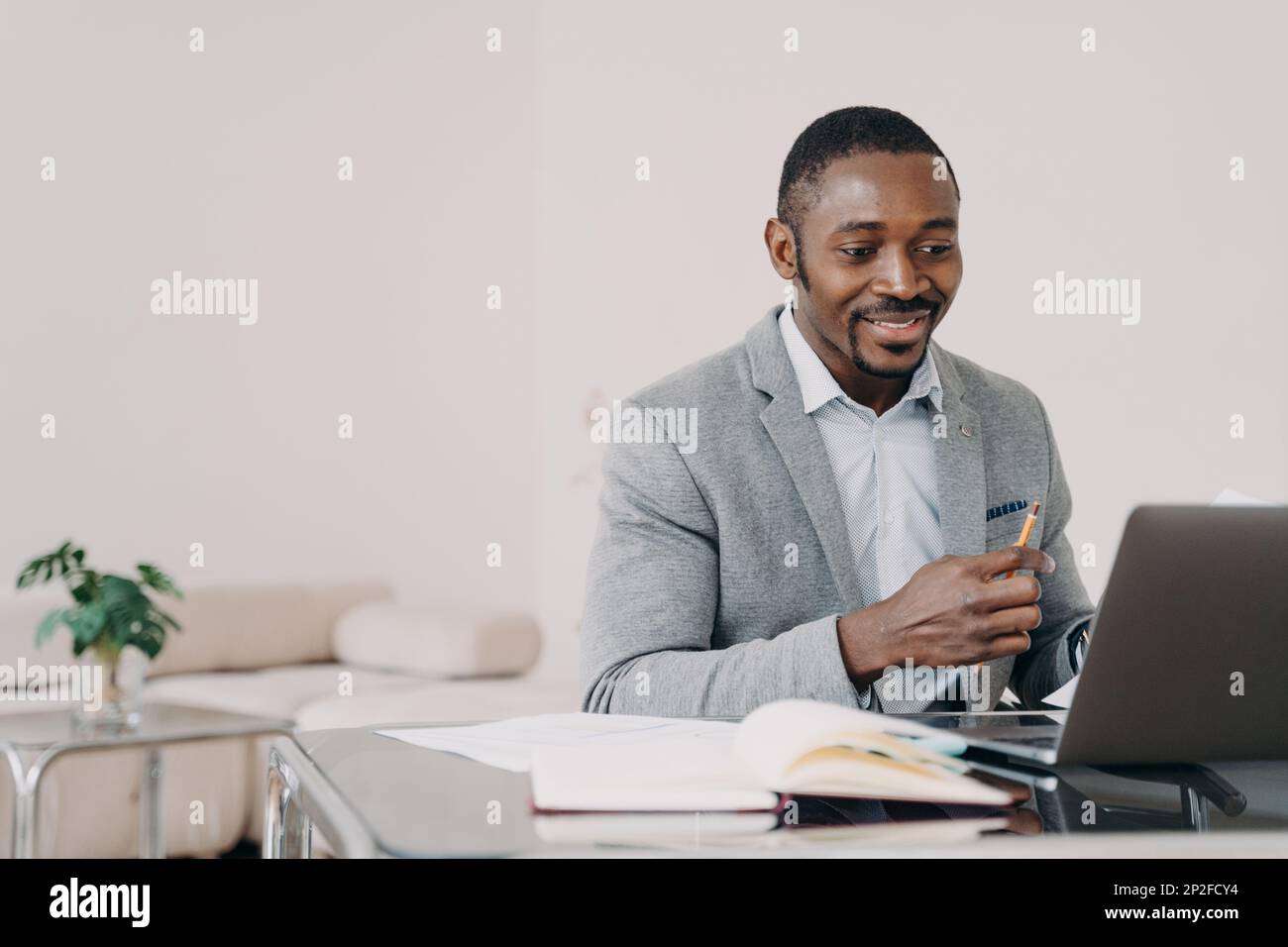 Lächelnder afroamerikanischer Mann, der online am Schreibtisch am Laptop arbeitet. Schwarzer männlicher Geschäftsmann, der auf einen Computerbildschirm schaut, E-Mails mit gutem ne liest Stockfoto