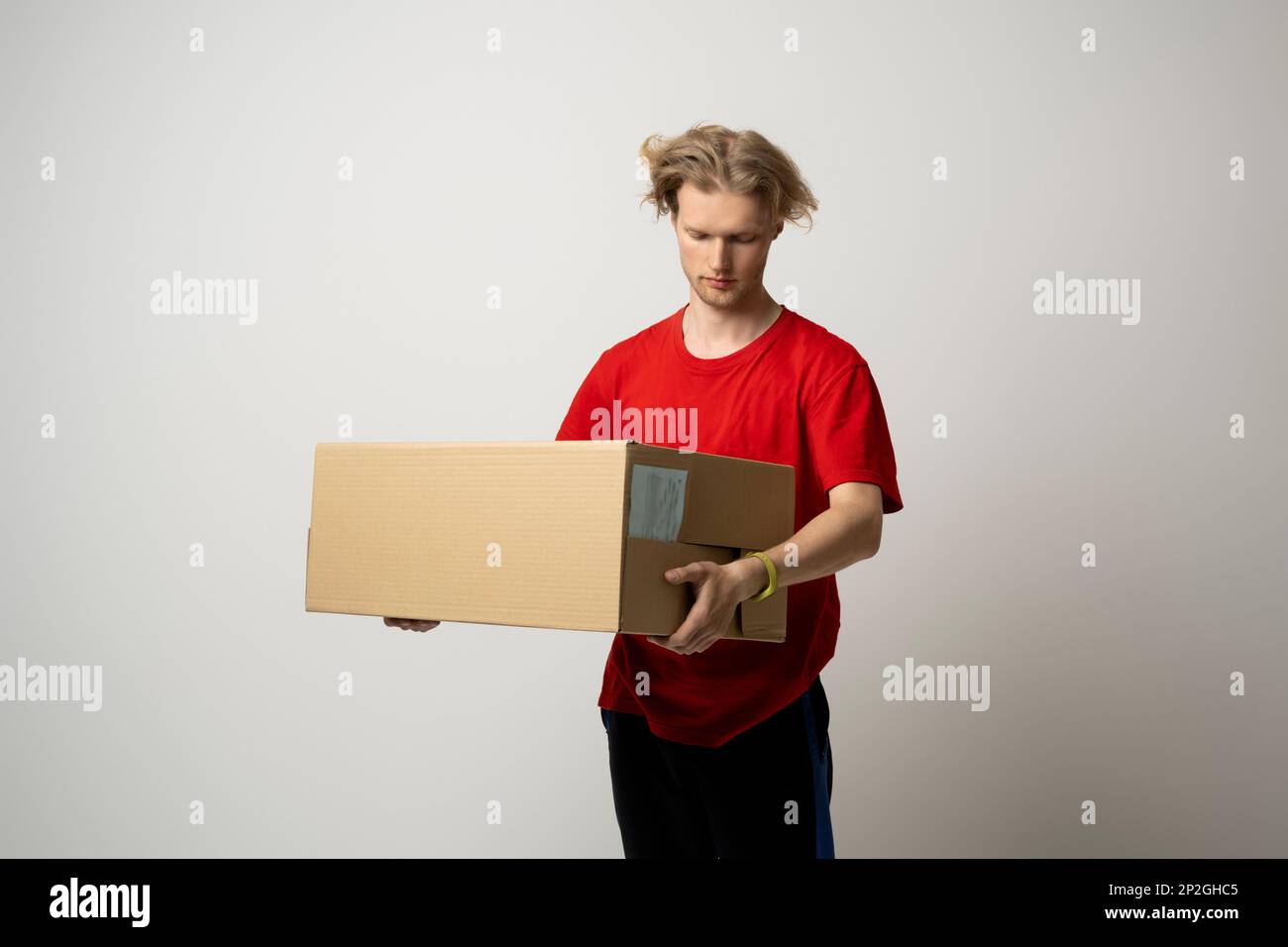 Der Zusteller in roter Uniform trägt einen Pappkarton in den Händen auf weißem Hintergrund. Der Zusteller übergibt die Paketlieferung. Stockfoto