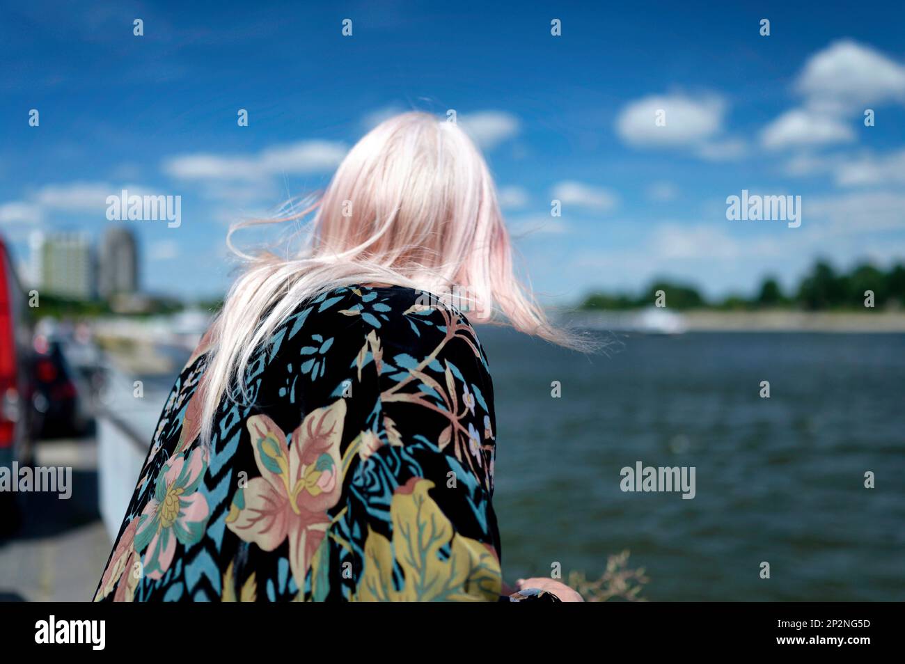 Eine Frau mit BlumenPoncho und rosa Haaren, die am Flussufer steht und nachdenklich in die Ferne blickt Stockfoto