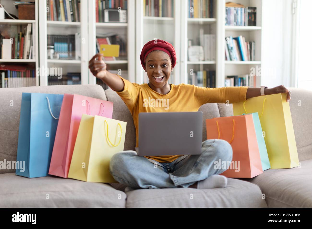 Eine glückliche schwarze Frau, die zwischen Einkäufen sitzt und ihre Kreditkarte vorzeigt Stockfoto