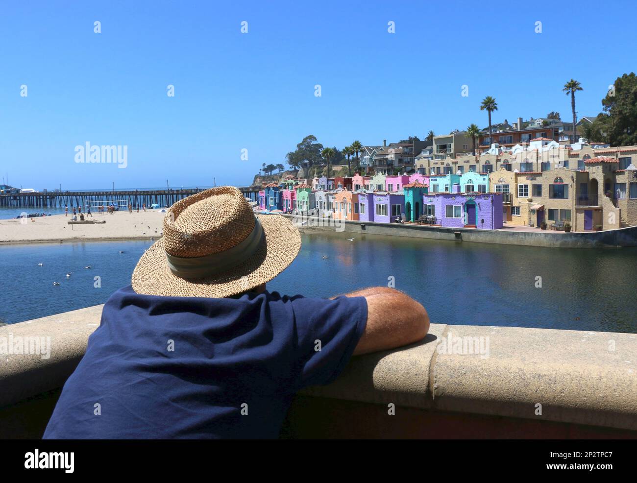 Ein älterer Mann mit Strohhut entspannt sich auf dem Balkon eines Restaurants mit Blick auf die bunten Hotelhäuser in Capitola, CA. Stockfoto