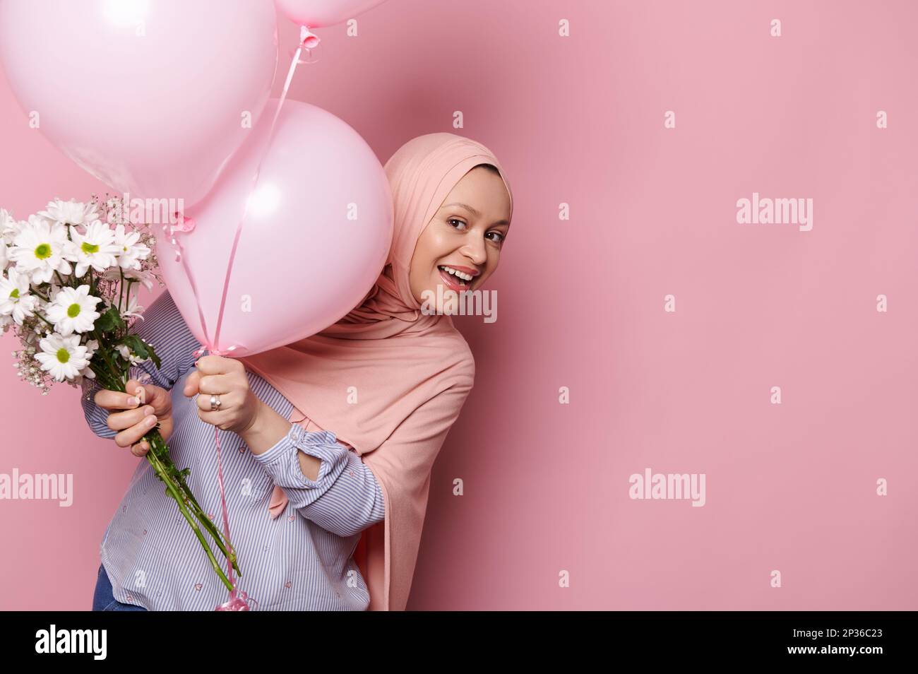 Eine wunderschöne muslimische Frau in pinkfarbenem Hijab und moderner Freizeitkleidung, die hinter pinkfarbenen Ballons aus dem Fenster guckt und vor der Kamera lächelt Stockfoto