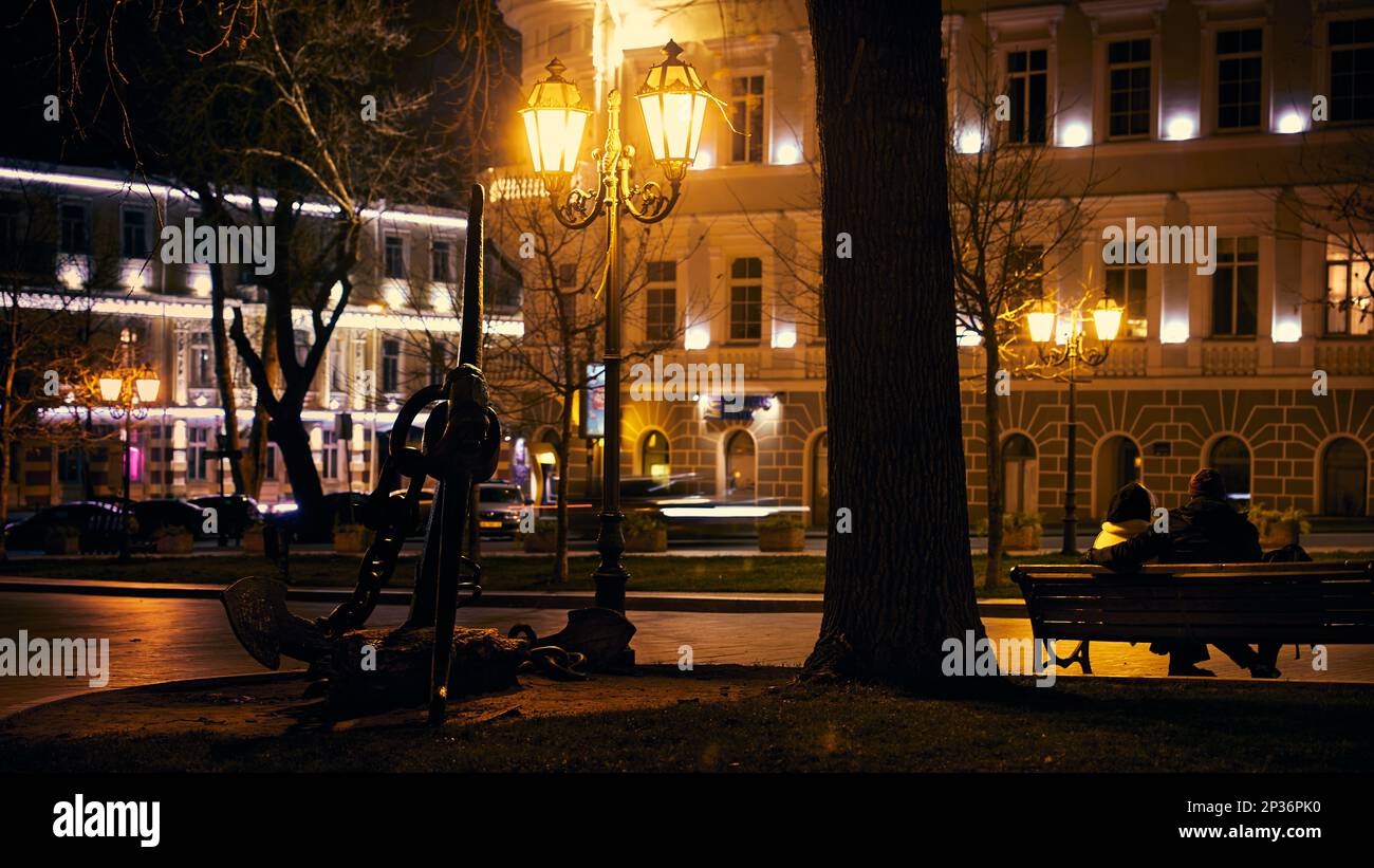 Ein Paar sitzt auf einer Bank im Licht der Straßenlaternen der Abendstadt und in der Nähe befindet sich ein Ankerdenkmal. Odessa Ukraine. Stockfoto